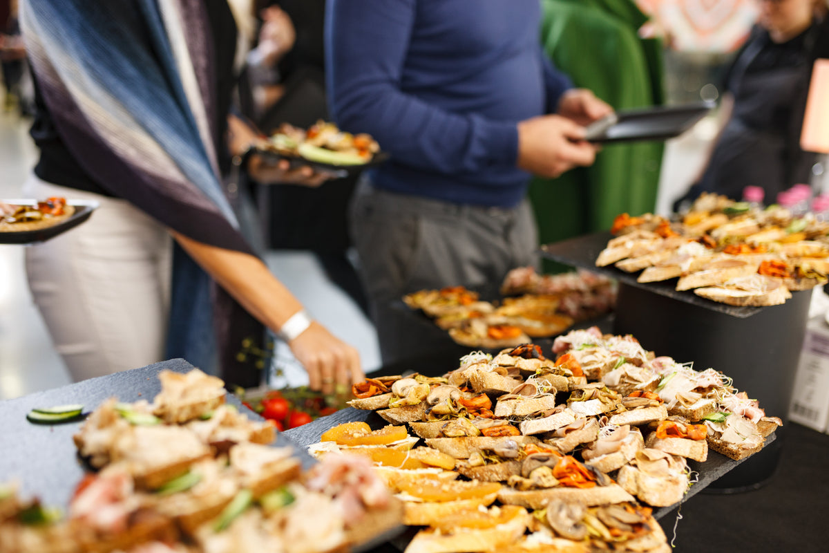 A catering buffet table filled with assorted sliced meats, cheeses, crackers, and garnishes, with several people standing in the background holding plates in a social event setting.