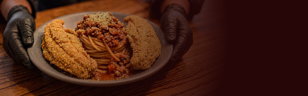 A plated pasta dish with meat sauce and breaded sides, held by Chef V. wearing black gloves, set on a wooden table.
